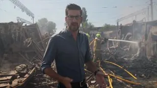 Dan Johnson wears a dark shirt and glasses, standing amongst rubble with smoke in the air and firefighers using a hose in the background