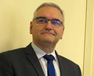 A man in a dark suit jacket, pale shirt and blue tie with short grey hair. He is wearing glasses. There is a plain cream-coloured wall behind him.