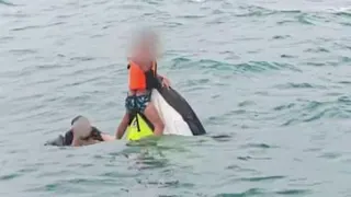 A boy wearing a lifejacket holds on to the top of a capsized jet ski as a man swims next to him in the sea