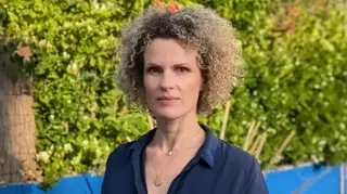 阿奈絲·德·沃斯穿著一件深藍色敞領襯衫，站在灌木叢和一堵低矮的亮藍色牆壁前。她戴著一條金項鍊，留著淺色捲髮。