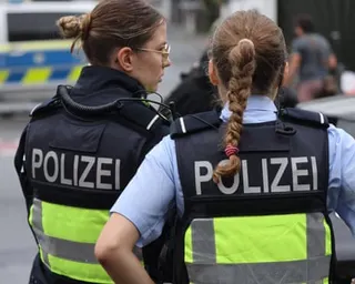 Two German police officers wearing "Polizei" vests stand together at a cordoned-off scene