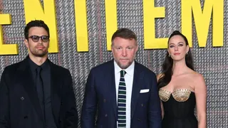 Theo James, Guy Ritchie and Kaya Scodelario pictured at the UK premiere of The Gentlemen in London in 2024. Guy Ritchie is dressed in a dark blue suit, white shirt and a tie featuring white stripes.
