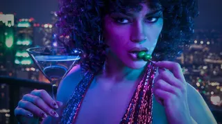 A pretty woman in a cocktail dress, with tanned skin and a loosely curled perm, holds what looks like a Martini cocktail and eats the olives on the stick seductively.
