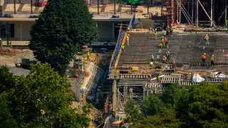 Construction workers in yellow and orange work on an in-progress project (President Trump's White House ballroom) near green trees.