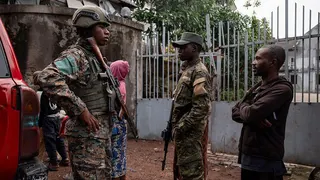 Two men dressed in military uniform and holding rifles stand alongside a member of the public in a brown hoody and trousers. A woman in a pink hoody has her back to the camera in the background