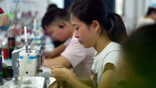 A woman sits at a sewing machine in a factory