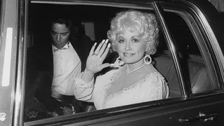 A woman is waving her hand as she sits in the backseat of a car with a man sitting next to her.
