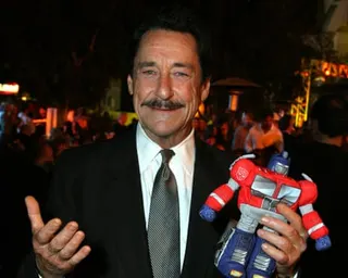Peter Cullen stands in a street in the dark holding a Transformers robot soft toy. He has dark slicked-back hair, wears a dark suit and tie, and has a debonair moustache.