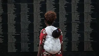 A woman with a baby on her back faces away from the camera. She is looking at a wall with names etched into it.