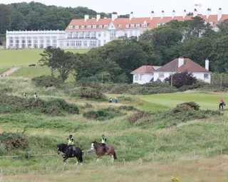 Mounted police patrol grassy dunes, with the course's large white hotel building behind. A golfer walking across a green is on the right
