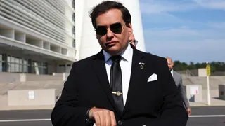 George Santos seen in a dark suit with a white shirt and pocket square and dark sunglasses. His expression is neutral.