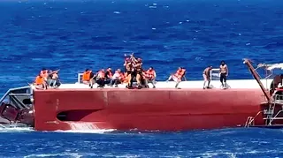 People wearing life jackets sit atop a capsized ferry as rescuers airlift one to safety.