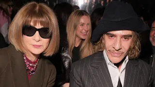 Anna Wintour in dark glasses and John Galliano in grey suit and black hat, seated together at a fashion show and looking expressionless at the camera