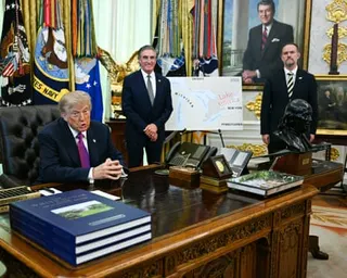a man speaks while seated a desk as two men stand near him