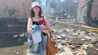 A young woman clutches a book to her chest and looks sad. Behind her is a bombed-out market structure. Destroyed books litter the ground around her