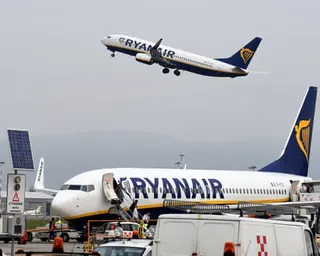 A Ryanair plane takes off overhead while another sits parked on the tarmac below