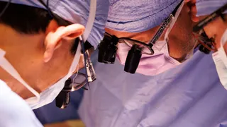 A tightly framed photograph shows several members of a surgical team gathered closely during a procedure. Each person is wearing blue surgical clothing, face masks, and magnifying lenses mounted on protective eyewear. The image focuses on their faces and equipment as they lean inward and look down toward an area outside the frame. Bright operating-room lighting illuminates the scene, while the background remains softly blurred in shades of blue. The composition emphasises concentration, precisio