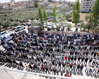 A high wide shot of hundreds of mourners standing in lines in an open square in the hilly village of al-Mughayyir