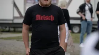 The torso of a man wearing a black t-shirt which says 'Arisch', German for Aryan written in a red Gothic-style font.