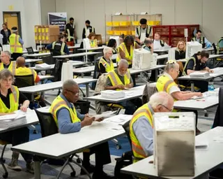 People wearing neon yellow vests sit one each at long tables sorting ballots.