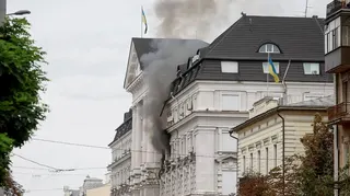Black smoke pours from two windows in a large government building with Ukrainian flags on the facades.